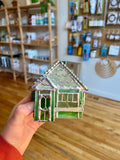 Hand holding a small green glass model of a house in a store setting with shelves and products in the background.