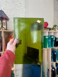 Person holding a green transparent piece of stained glass in front of a blurred interior. 