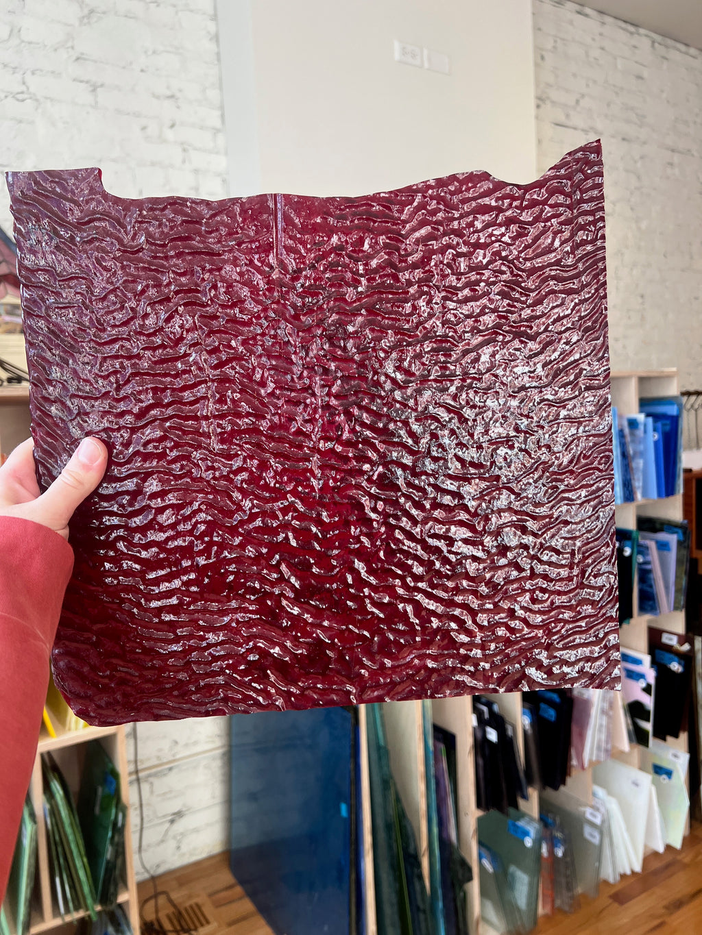 Person holding a red textured piece of stained glass in front of a blurred sunny interior. 