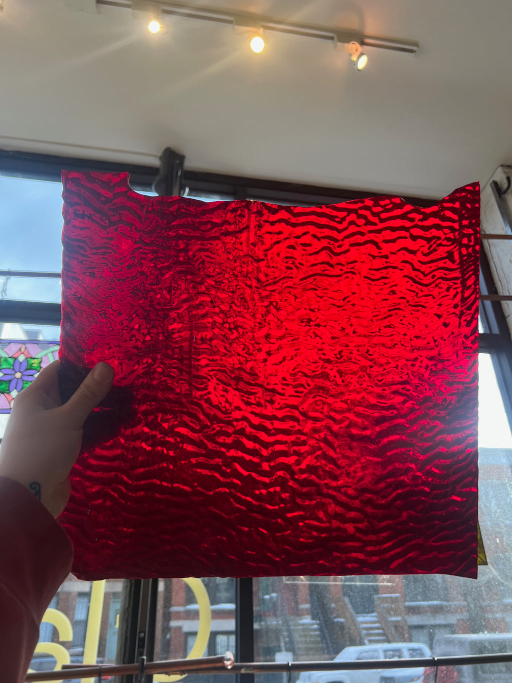 Person holding a red textured piece of stained glass in front of a blurred sunny window. 