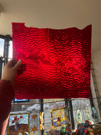 Person holding a red textured piece of stained glass in front of a blurred sunny window. 