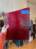 Person holding a red textured piece of stained glass in front of a blurred interior. 