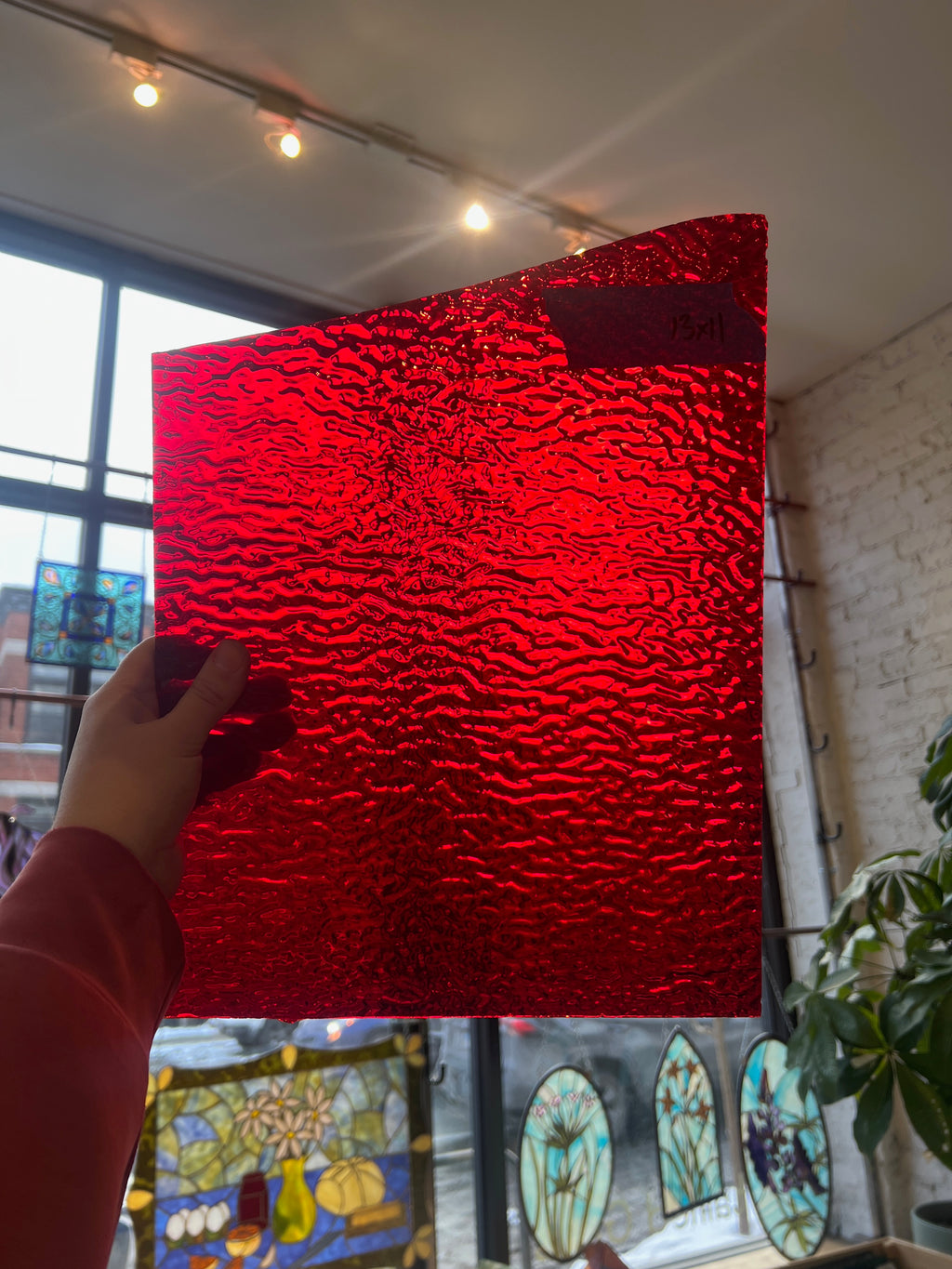 Person holding a red textured piece of stained glass in front of a blurred sunny window. 