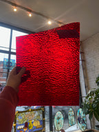 Person holding a red textured piece of stained glass in front of a blurred sunny window. 